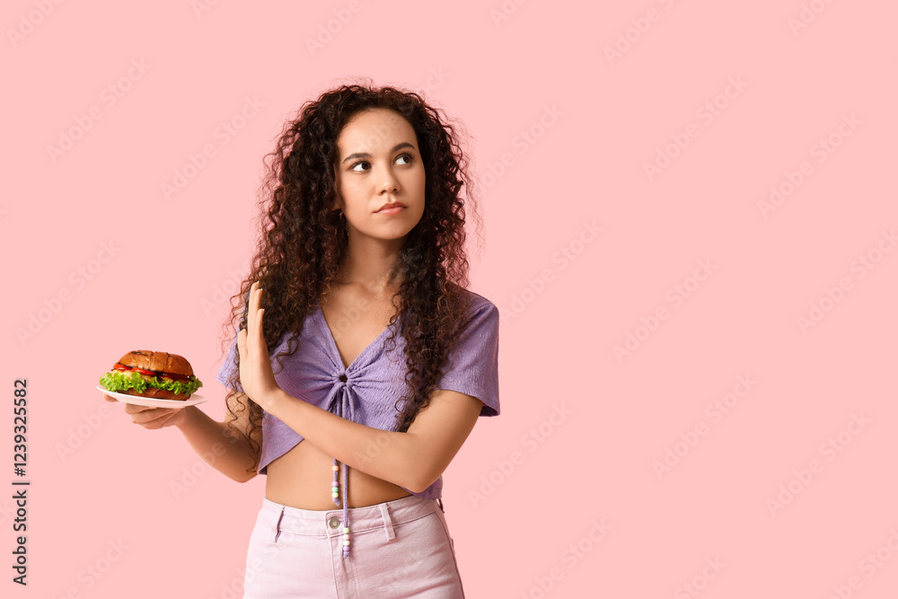 Young African-American woman showing stop gesture against burger on pink background. Diet concept Young African-American woman showing stop gesture against burger on pink background. Diet concept