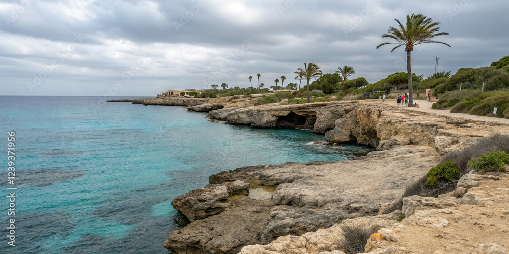 A rocky shoreline features turquoise waters lapping against cliffs, dotted with palm trees. Visitors stroll along the coast under a partly cloudy sky, enjoying the tranquil setting A rocky shoreline features turquoise waters lapping against cliffs, dotted with palm trees. Visitors stroll along the coast under a partly cloudy sky, enjoying the tranquil setting