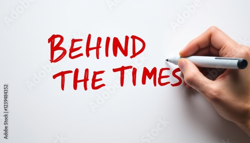 A hand scribbling 'behind the times' on a white surface with a red marker