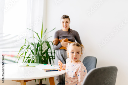 Girl sits at table, observing mom with newborn. Minimalist interior, natural light, moments of older child adapting to new sibling. Real family life, unposed and authentic. photo