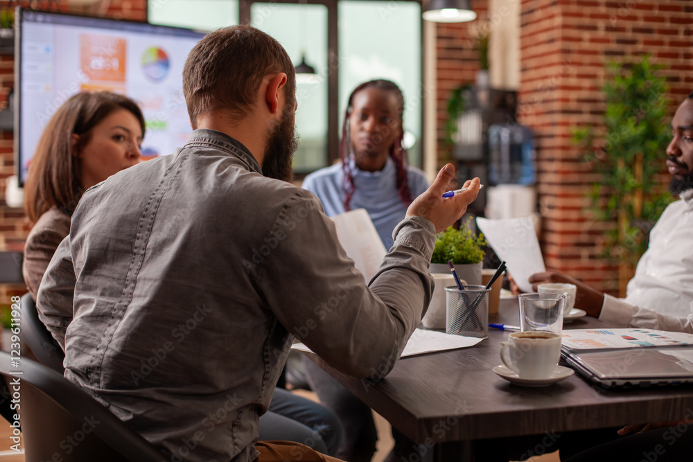 Rearview of caucasian man providing updates of business project to his colleagues in office. Multiethnic coworkers listening to white businessman during meeting in brick wall workspace. Rearview of caucasian man providing updates of business project to his colleagues in office. Multiethnic coworkers listening to white businessman during meeting in brick wall workspace.