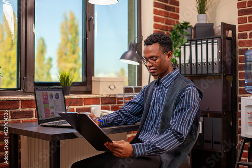 African American professional, sitting at desk near window, reviewing financial paperwork and marketing strategy ideas. Manager with clipboard reading project development plans in brick wall office.