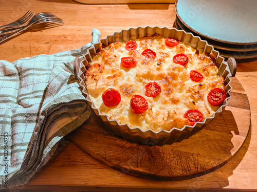 -brown casserole topped with cherry tomatoes, resting on a wooden board with a plaid cloth and cutlery nearby. photo