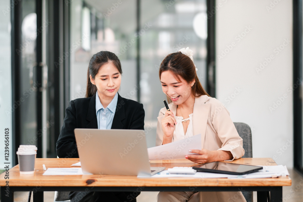 Two businesswomen working together at a desk in a contemporary office, discussing documents and using a laptop. Two businesswomen working together at a desk in a contemporary office, discussing documents and using a laptop.