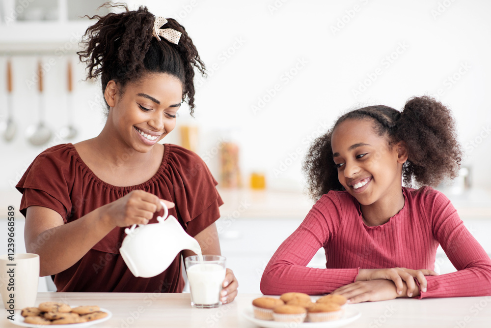 Pretty black mother pouring fresh milk for her teen daughter, happy afro-american girl having breakfast with her loving mom, kitchen interior. Healthy diet, calcium, vitamins in kids food concept