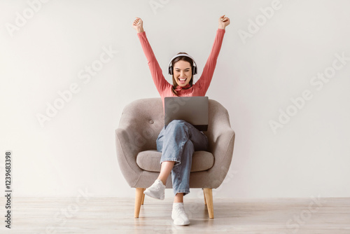 Wall Mural Full length of excited young lady with laptop and headphones raising her arms up