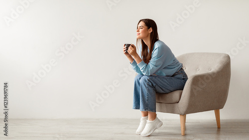 Time for coffee. Charming millennial woman sitting in armchair, enjoying cup of hot drink against white studio wall, banner with free space. Young lady relaxing with mug of warm beverage photo