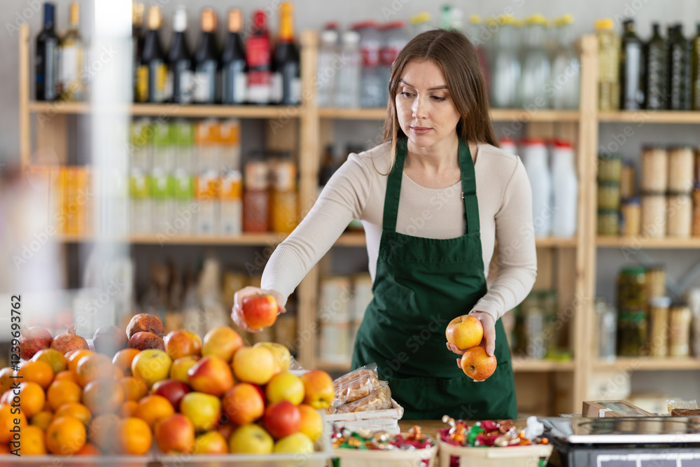 Friendly sales girl works at grocery store. She puts apples in window, replenishes fruit stocks on counter, corrects appearance of product display Friendly sales girl works at grocery store. She puts apples in window, replenishes fruit stocks on counter, corrects appearance of product display
