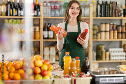 Polite young female seller is kindly offering bottles of natural juice in grocery store photo