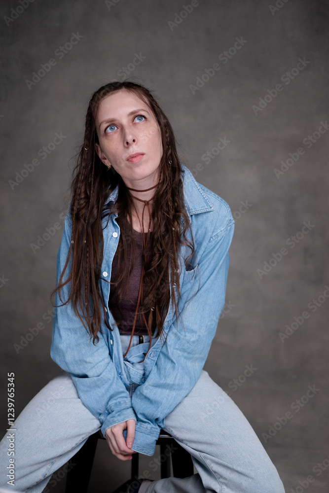 a beautiful and long-haired woman with big blue eyes in stylish clothes posing in a photo studio. the model sits on a chair and poses in denim clothes on a gray photo background. a beautiful and long-haired woman with big blue eyes in stylish clothes posing in a photo studio. the model sits on a chair and poses in denim clothes on a gray photo background.