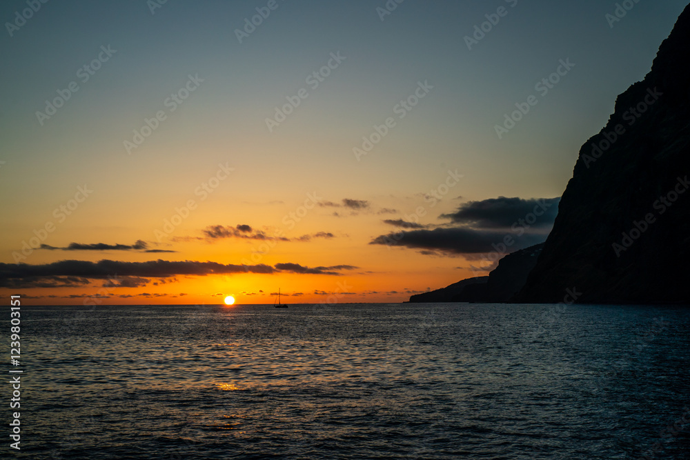 Serene sunset over the ocean, silhouetted cliff and a distant sailboat creating a picturesque view. Serene sunset over the ocean, silhouetted cliff and a distant sailboat creating a picturesque view.
