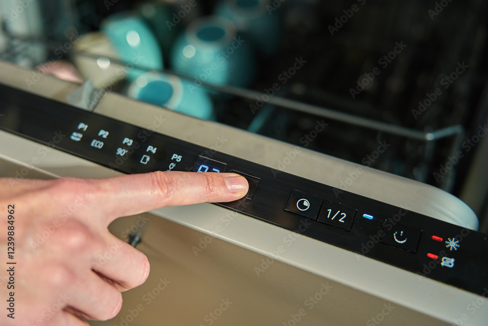 Close-up of female finger pressing button on dishwasher control panel. Woman using dishwasher in modern kitchen. Modern kitchen appliance with digital settings for efficient dishwashing