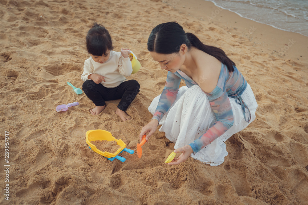happy toddler baby girl playing sand toy with mother on sea beach in Pattaya, Thailand happy toddler baby girl playing sand toy with mother on sea beach in Pattaya, Thailand