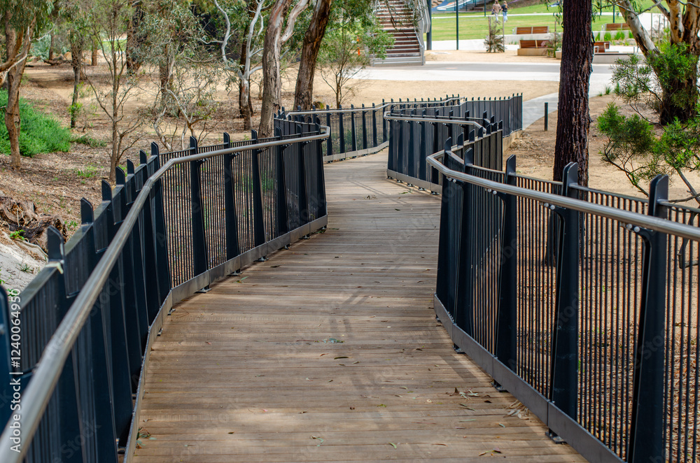 A curved wooden boardwalk with metal safety railings winds through an Australian park, providing an accessible route through public parkland—an inclusive community amenity. A curved wooden boardwalk with metal safety railings winds through an Australian park, providing an accessible route through public parkland—an inclusive community amenity.