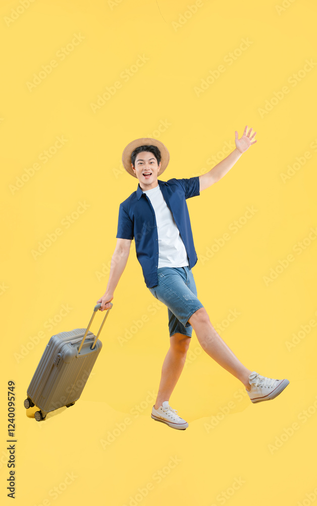 Full body image of a young Asian man pulling a suitcase and posing on a yellow background
