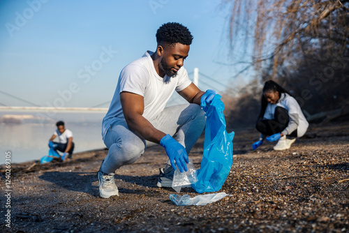 Happy multicultural volunteer putting plastic and paper waste into bag and cleaning riverbank with his eco team. photo