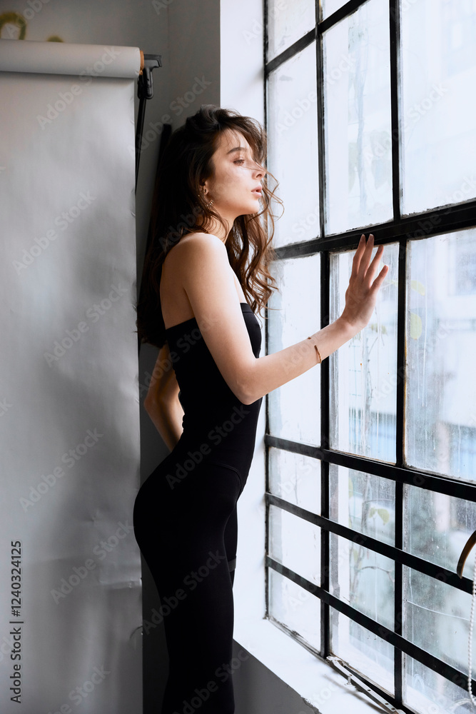 Fototapeta premium A woman in a black dress is standing in front of a window. She is wearing a gold necklace and a gold bracelet. The image has a mood of elegance and sophistication