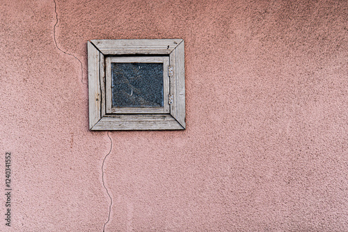 A small, weathered wooden window with a cracked glass pane is set into a textured pink stucco wall. The aged frame, with its rough, faded wood, stands out against the soft, muted pink backdrop. 
