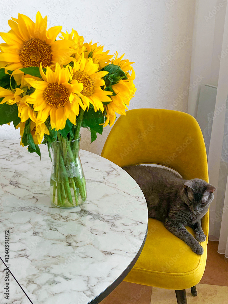 A grey cat is lying on a yellow armchair near a table with a bouquet of sunflowers