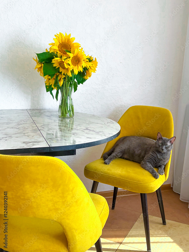 A grey cat is lying on a yellow armchair near a table with a bouquet of sunflowers