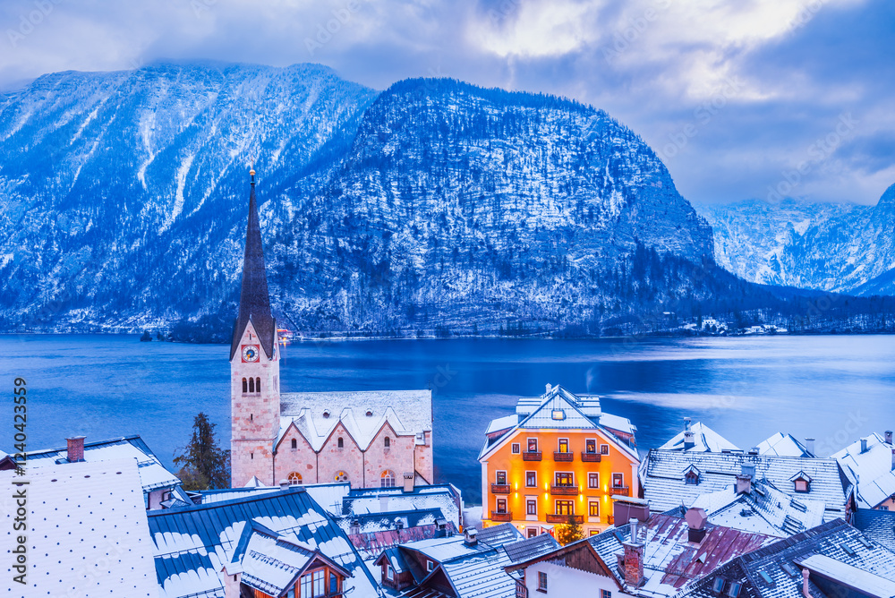 Hallstatt, Austria - Winter snowy twilight of tranquil Alpine village, snow-covered rooftops, serene lake, majestic mountain ridge.