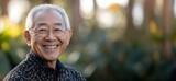 Smiling elderly Asian man with glasses in nature, showcasing joy and wisdom, wearing traditional patterned shirt, soft focus background, copy space for text