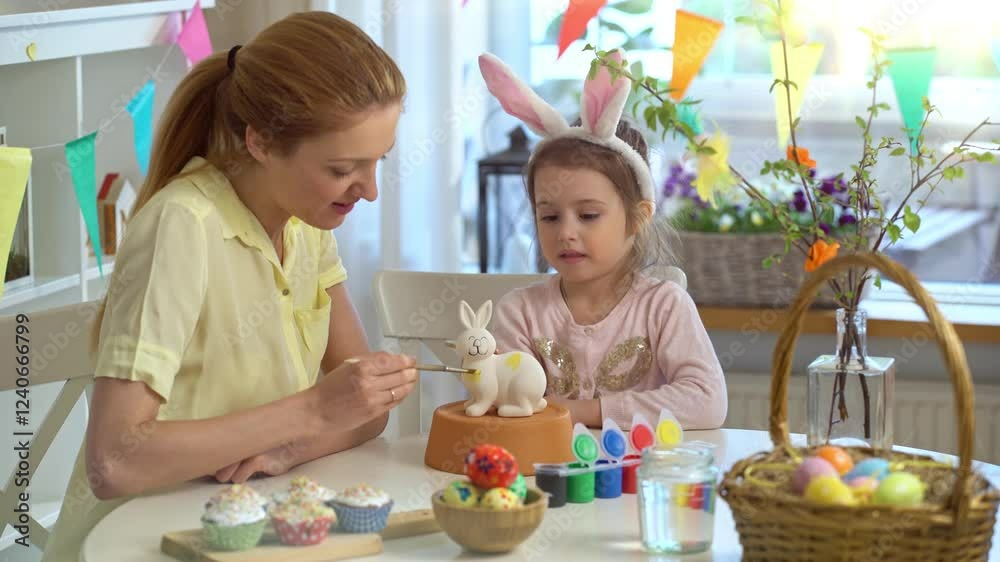 Mother and daughter paint a ceramic bunny together for Easter. The girl, wearing bunny ears, carefully applies yellow paint while her mother assists. The table is filled with colorful paints Mother and daughter paint a ceramic bunny together for Easter. The girl, wearing bunny ears, carefully applies yellow paint while her mother assists. The table is filled with colorful paints