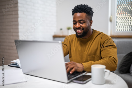 Happy young black businessman using laptop while enjoying working at his cozy living room.