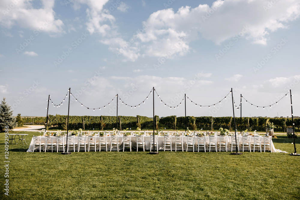 Fototapeta premium Elegant outdoor dining setting with a long banquet table, white chairs, fairy lights, lush greenery, and a vineyard backdrop.