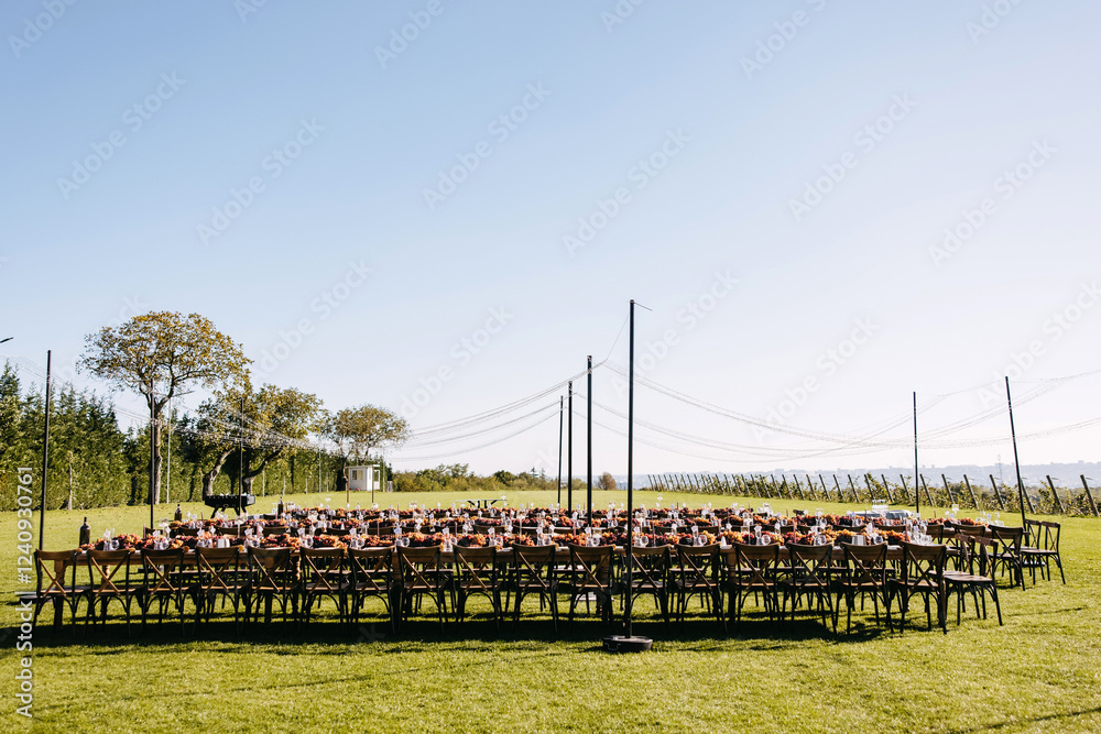 Fototapeta premium An elegant outdoor wedding reception setup on a lush green lawn with long tables, wooden chairs, and warm floral decorations under string lights