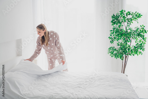 Young woman in cozy pajamas tidying up bed in bright bedroom with natural daylight