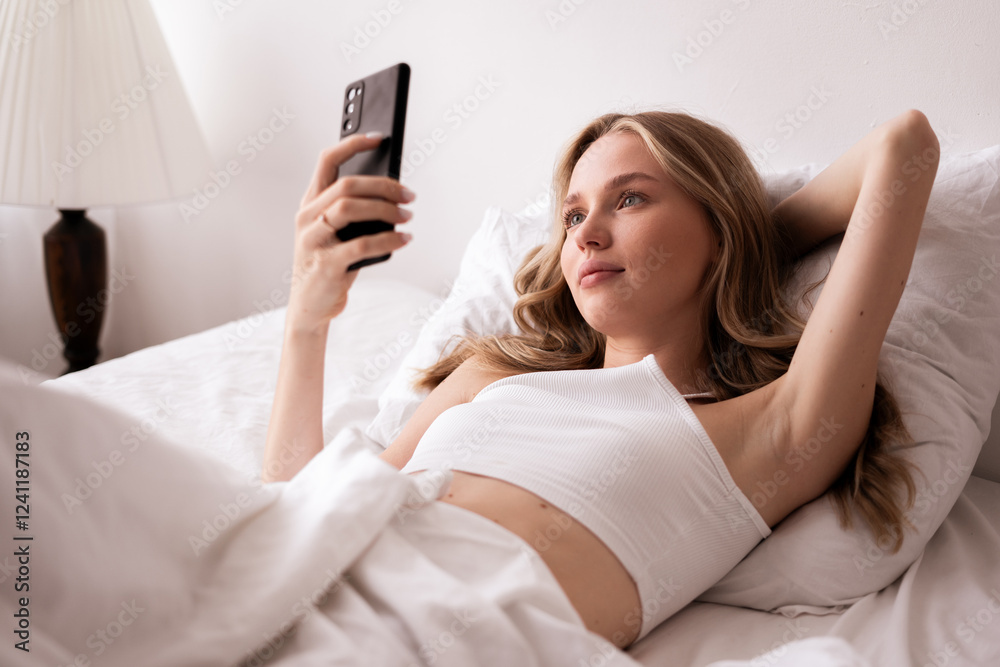 Young woman with long hair using smartphone lying in bed in the morning enjoying weekend relaxation Young woman with long hair using smartphone lying in bed in the morning enjoying weekend relaxation
