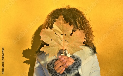 Chica joven muy abrigada que se cubre totalmente el rostro con una hoja de arbol que lleva pintada un emoticono de infeliz. photo