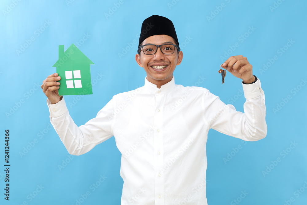 Asian muslim man holding small paper house , posing over blue color background wall in studio with copy space for promotion content or design. Asian muslim man holding small paper house , posing over blue color background wall in studio with copy space for promotion content or design.