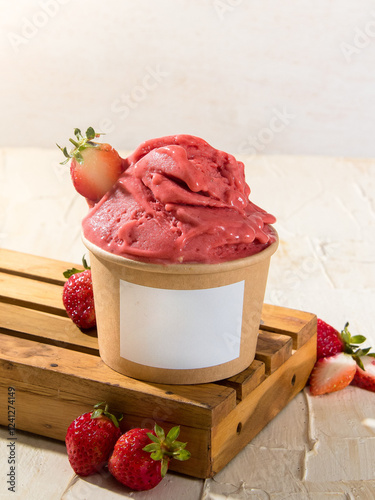 Vibrant strawberry gelato sits in a paper cup surrounded by fresh strawberries on a rustic wooden surface showcasing delightful summer flavors