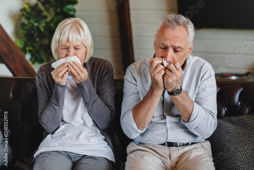 Virus and flu concept. Mature family sitting on sofa and blowing their noses. Sick couple are trying to sneeze in the napkin. They caught a cold and now have to take some medicine to get better. photo