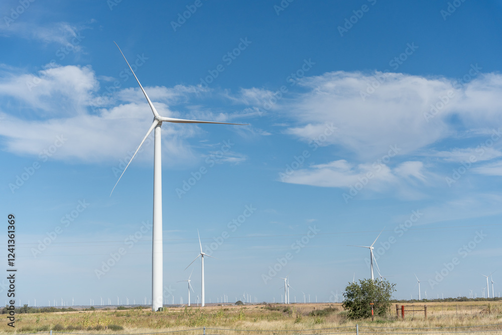 Vast field with a beautiful blue sky above it. In the distance, there are several wind turbines, which are spread out across the field. The wind turbines are of different sizes Vast field with a beautiful blue sky above it. In the distance, there are several wind turbines, which are spread out across the field. The wind turbines are of different sizes