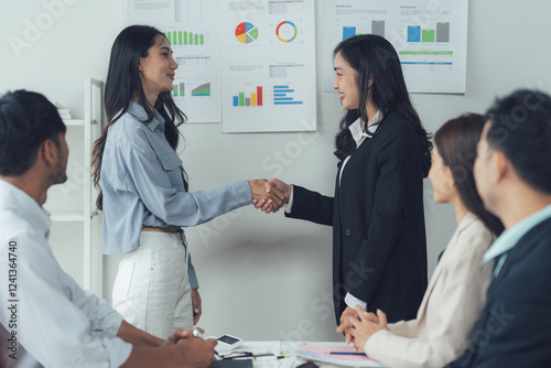 Business Deal Handshake: Two businesswomen seal a deal with a firm handshake in a modern office setting, surrounded by their colleagues. The image symbolizes partnership, collaboration. photo