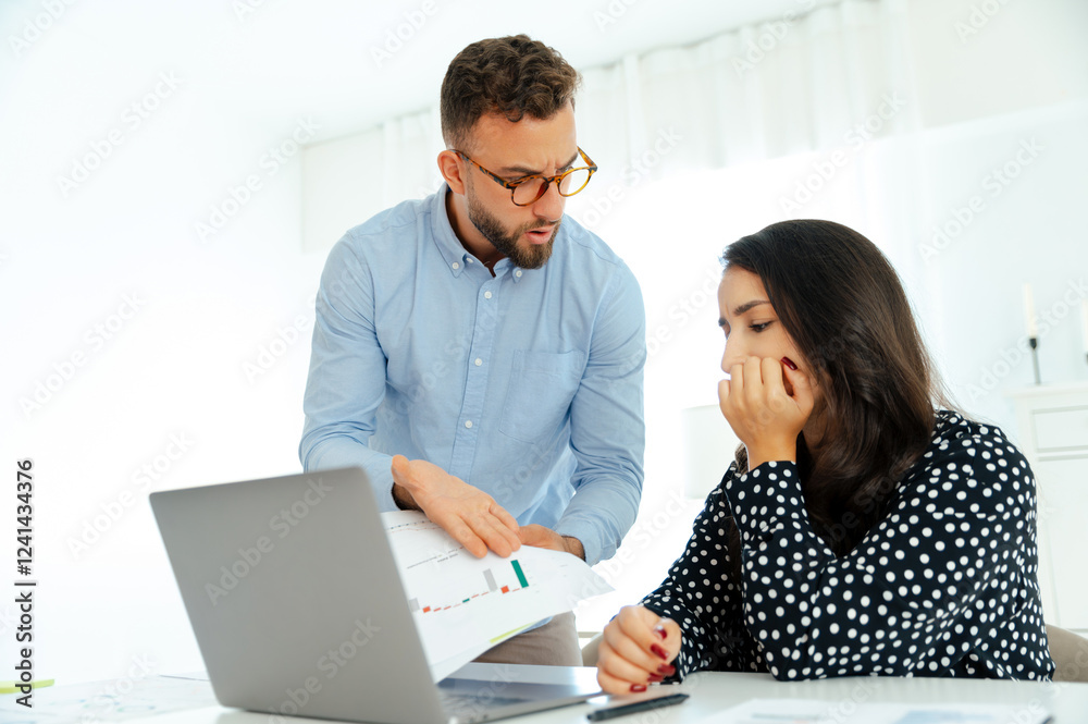 Misunderstanding between coworkers. A worried man in glasses and a blue shirt shows financial chart to his female colleague, both looking at a graph with concerned expressions, analyzing reports Misunderstanding between coworkers. A worried man in glasses and a blue shirt shows financial chart to his female colleague, both looking at a graph with concerned expressions, analyzing reports
