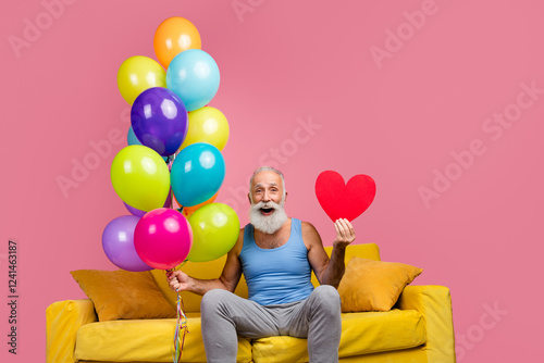 Cheerful senior man on sofa with balloons and red heart against pink background photo