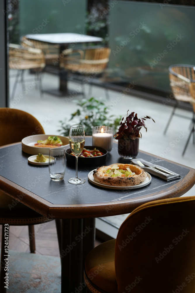 Fototapeta premium A restaurant table with a pizza, a glass of white wine, and two plates with other food on it. The table is set for two person dinner