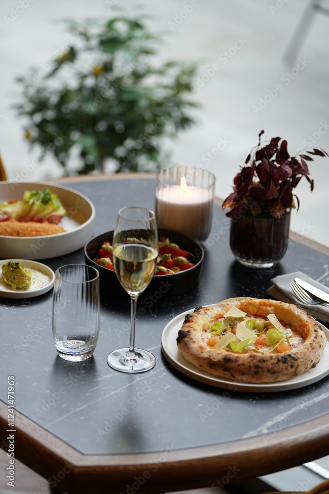 A restaurant table with a pizza, a glass of white wine, and two plates with other food on it. The table is set for two person dinner A restaurant table with a pizza, a glass of white wine, and two plates with other food on it. The table is set for two person dinner