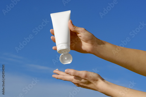 Female hands squeezes a white mock-up tube of cream on her finger on a blue sky background. Concept of natural products for body and face care. Image for your design