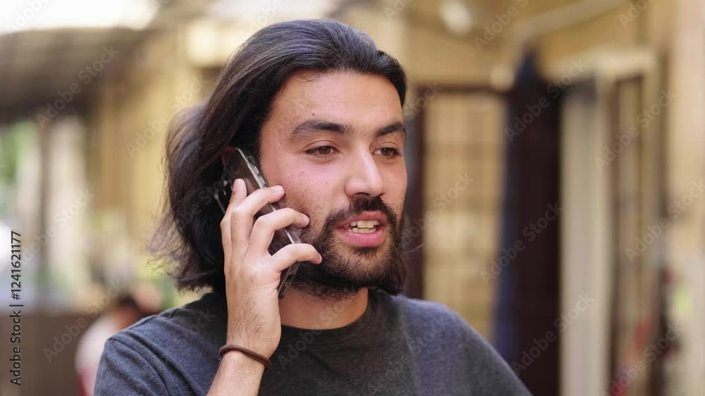 A young man with long hair and a short beard stands while talking on the phone, appearing engaged in the conversation. A young man with long hair and a short beard stands while talking on the phone, appearing engaged in the conversation.