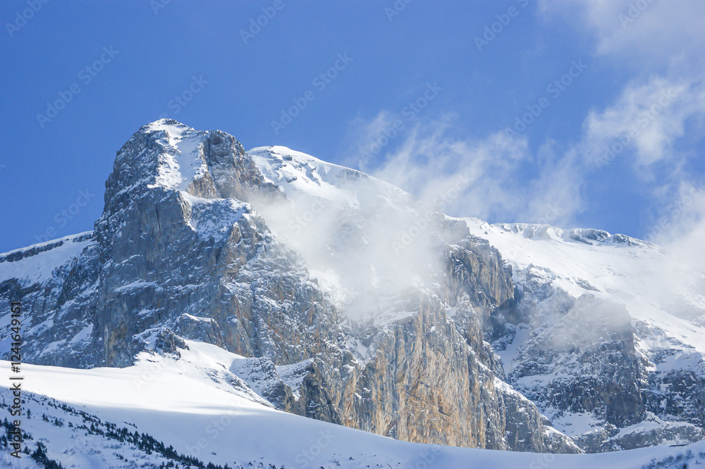 Naklejka premium A snowy rocky mountain peak with wind blowing snow and a blue sky near a ski resort in the French or Swiss Alps in the winter