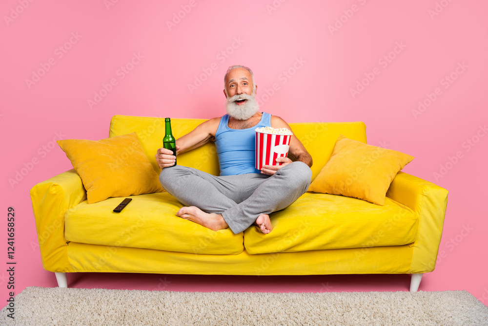 Obraz premium Elderly man relaxing on a vibrant yellow sofa with popcorn and drink against pink background