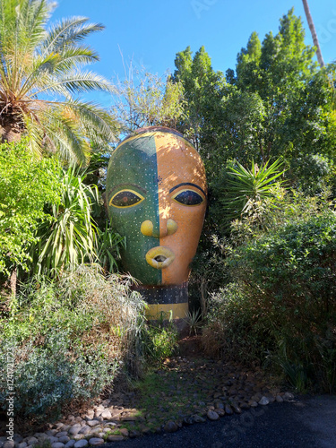 Unique Large Artistic Mask Surrounded by Lush Greenery in a Vibrant Garden Setting