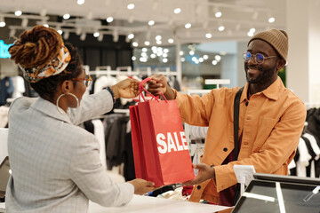 Happy young male consumer passing red sale paperbags to clerk over counter before paying for new attire in boutique