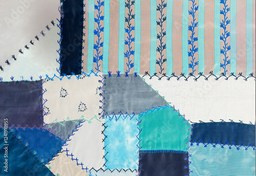 Old sample fabric texture background shows thread stitches in fancy patterns joining geometric forms together in shades of aqua, neutral and blue . Material is retro.