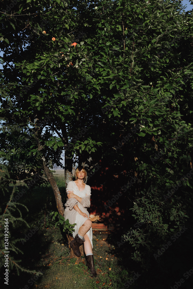 Naklejka premium A young, pretty woman in a beige dress is standing in a garden surrounded by plants. A blonde model is posing in a creative photo shoot in a rural style.